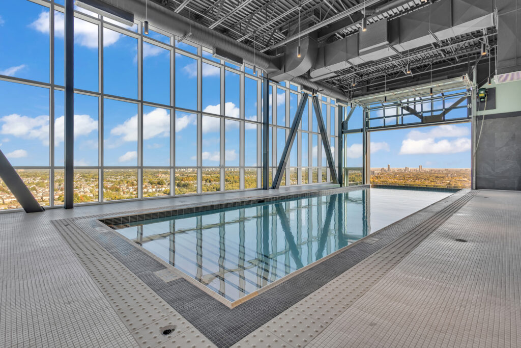 Indoor swimming pool with clear blue water, surrounded by tall windows and a spacious building structure.