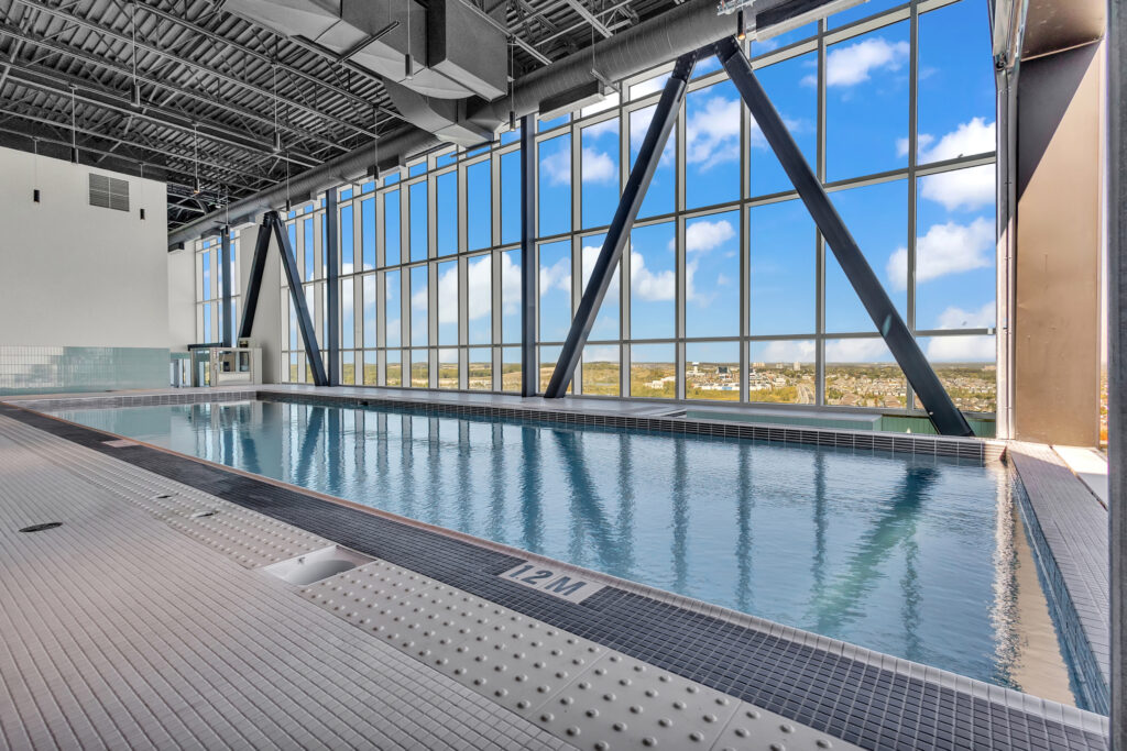 Indoor swimming pool with clear blue water, surrounded by tall windows and a spacious building structure.
