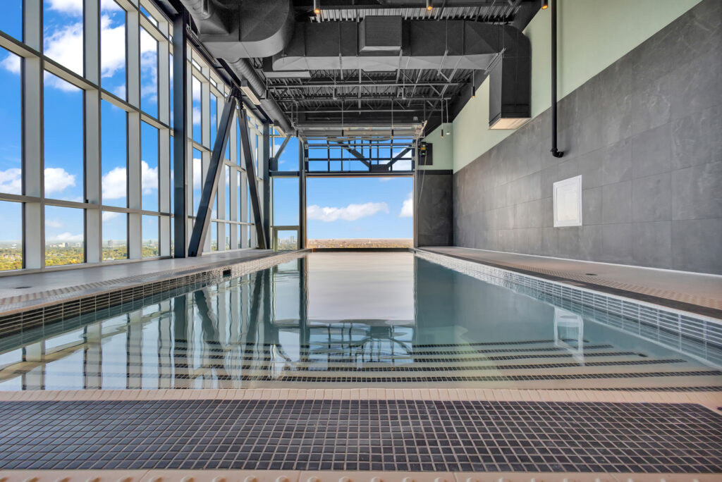 Indoor swimming pool with clear blue water, surrounded by tall windows and a spacious building structure.